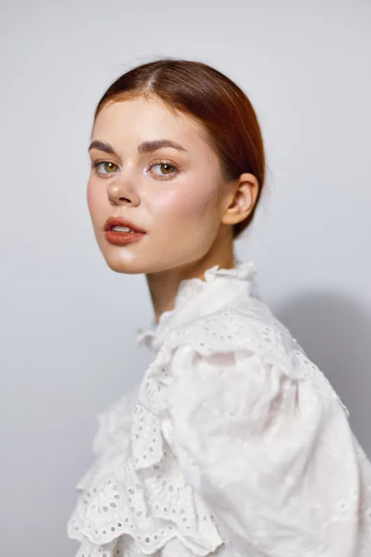 Elegant young woman with a serene expression, wearing a romantic white lace blouse, capturing a timeless beauty in a soft pastel background