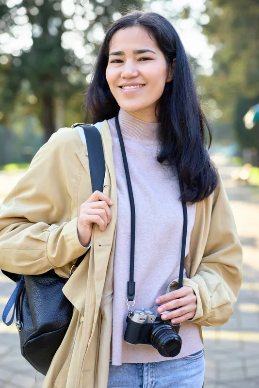 Woman with camera and backpack smiling outdoors, casual jacket, daylight photography vibe capturing everyday moments and travel curiosity