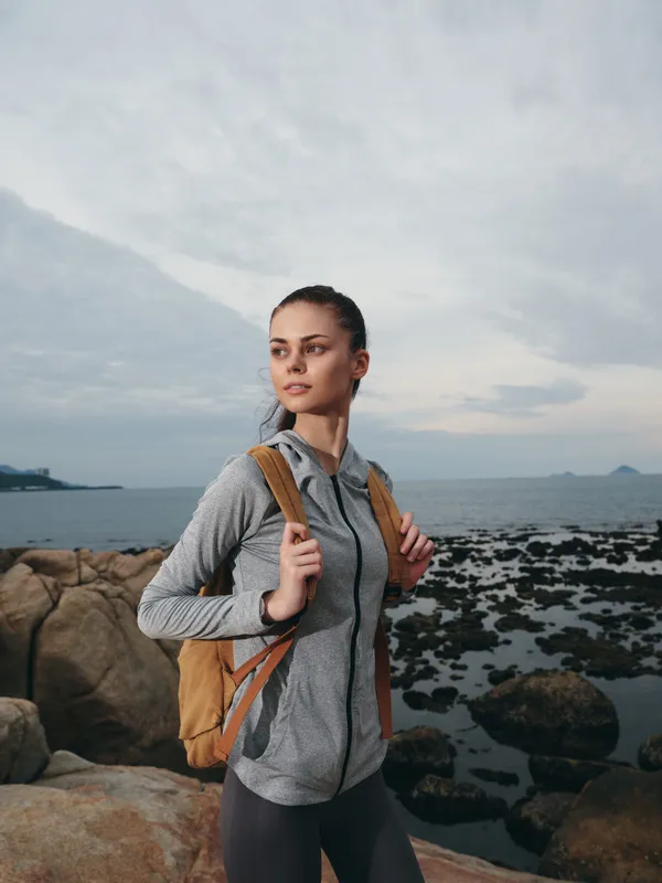 Young woman hiking by the sea, wearing sporty clothes and a backpack, showcasing a determined expression with a cloudy sky in the background