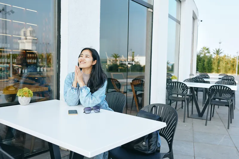 Woman at an outdoor cafe table, smiling with daylight glow, sunglasses on the table, glass storefronts in the background, relaxed mood, casual denim style in warm sunlight