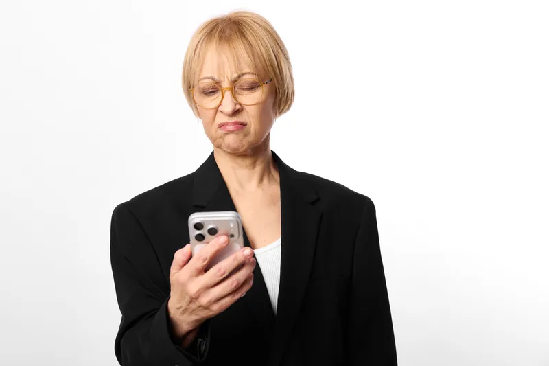 Woman smartphone suit glasses studio upset mature expression, holding a phone with both hands, soft light, white background, business communication, isolated