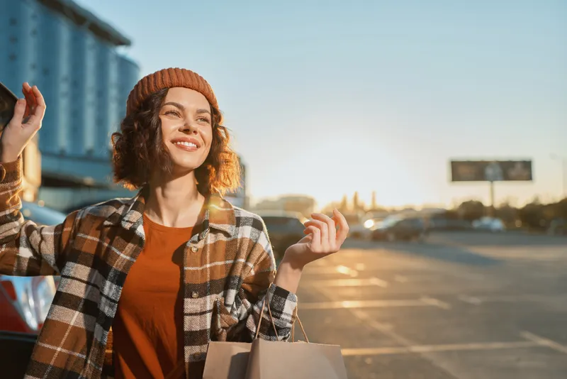Woman smiling with shopping bags in an urban street at golden hour sunlight, candid lifestyle portrait conveying authenticity, mindful living and emotional storytelling in autumn glow.