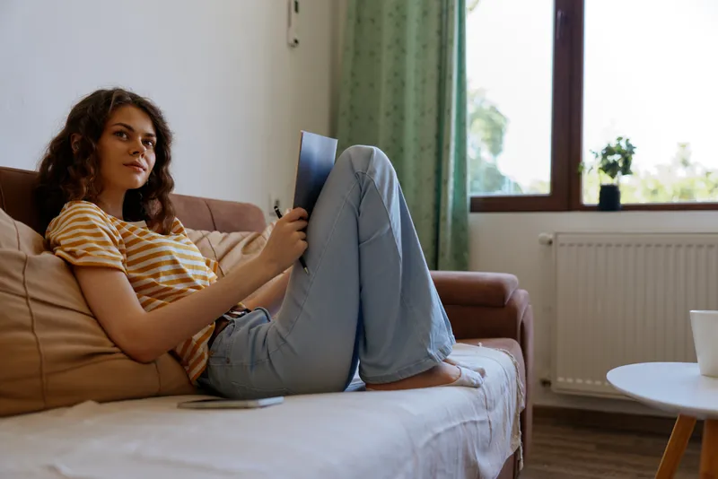 Woman relaxing at home sits on a sofa with a laptop, casual striped tee, light jeans, soft natural light, comfortable workspace ambiance, home life and easy mood showcased.