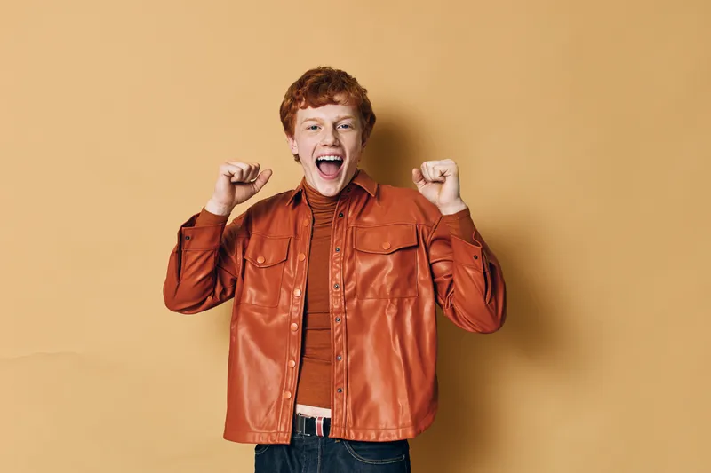 Young man with red hair smiling and celebrating with raised fists wearing brown leather jacket and orange shirt against beige background. Happy excited person expression.