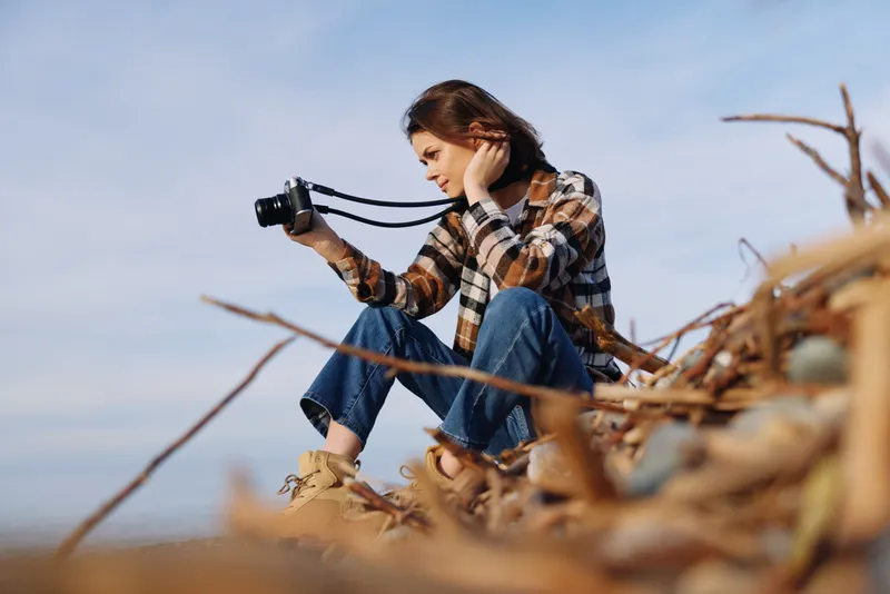 Photographer, woman and camera in an outdoor setting with natural light, casual plaid shirt, sitting on driftwood, capturing landscape moments with focused expression and calm mood.