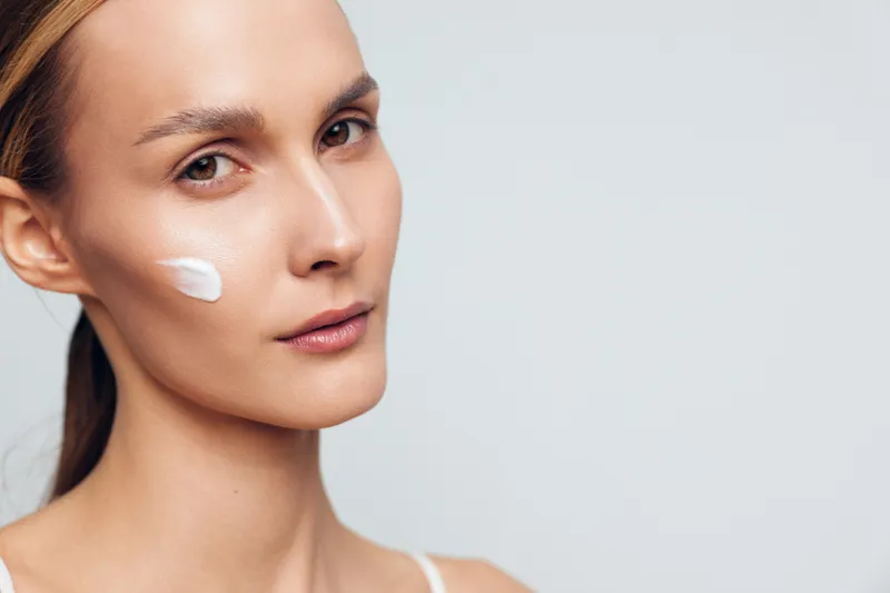 A young woman with clear skin applies white face cream on her cheek, looking at the camera. The minimalistic background highlights skincare and natural beauty concepts.