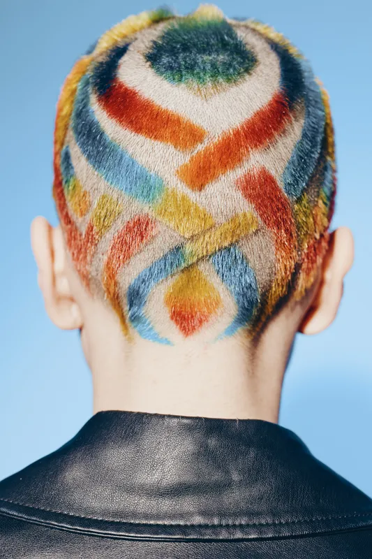 Woman with colorful shaved head hair design featuring abstract patterns, bright colors, and a modern edgy style against a soft blue background.