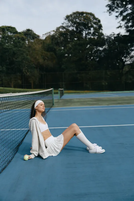 Young woman in sportswear sitting on a tennis court, displaying a calm emotion while wearing a white outfit and headband, with a focused background of greenery