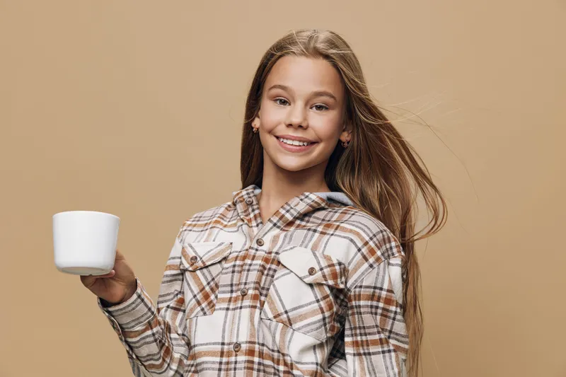 Girl smiling holding cup wearing plaid shirt with long hair smiling at camera on beige background. Youth, beauty and casual style with happiness and relaxed expression.