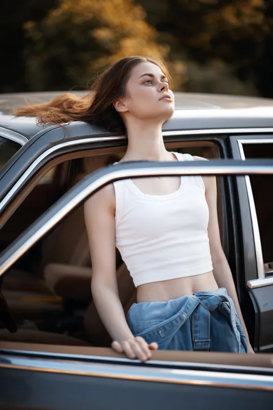 A young woman enjoying a sunny day, wearing a white crop top and denim jeans, leaning out of a vintage car with a relaxed expression, surrounded by a warm autumn background.