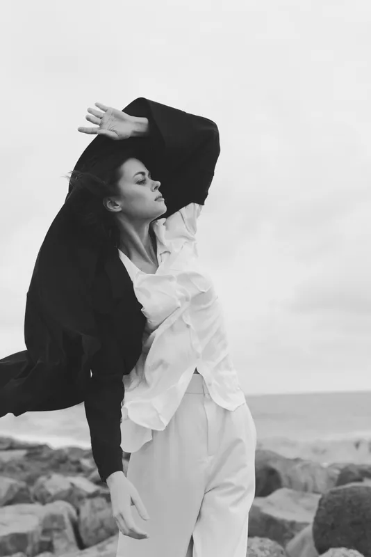 Fashionable woman on a windy beach, coat billowing, white shirt fluttering, graceful movement by the sea in monochrome tones.