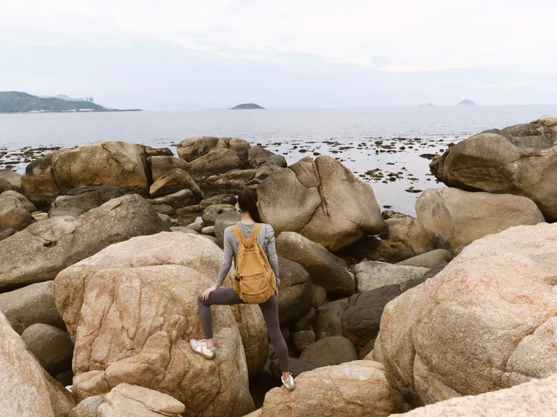 Young woman exploring rocky coastline in activewear, looking towards a tranquil sea, showcasing adventure and outdoor lifestyle with a sense of freedom
