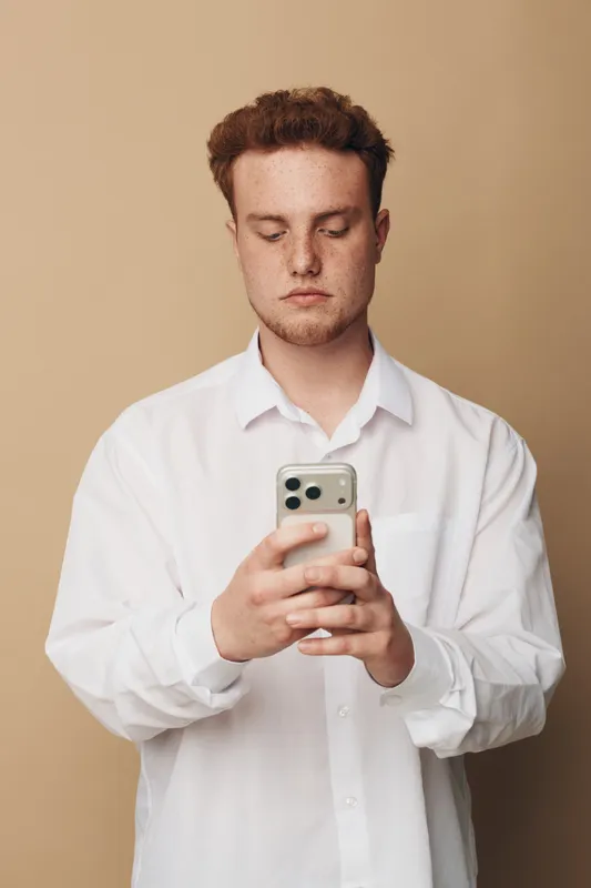 Young man with red hair and freckles wearing white shirt looks at smartphone with serious expression on beige background. Concept of technology, communication, and modern lifestyle.