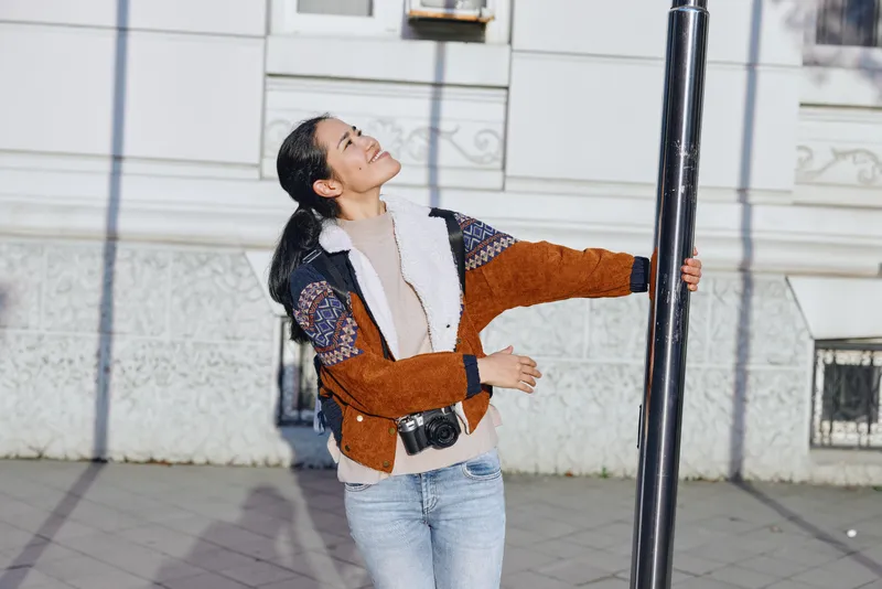 Street scene with a joyful woman hugging a lamp post in an outdoor urban setting, denim jeans, textured jacket, bright light, dynamic pose, candid street style moment