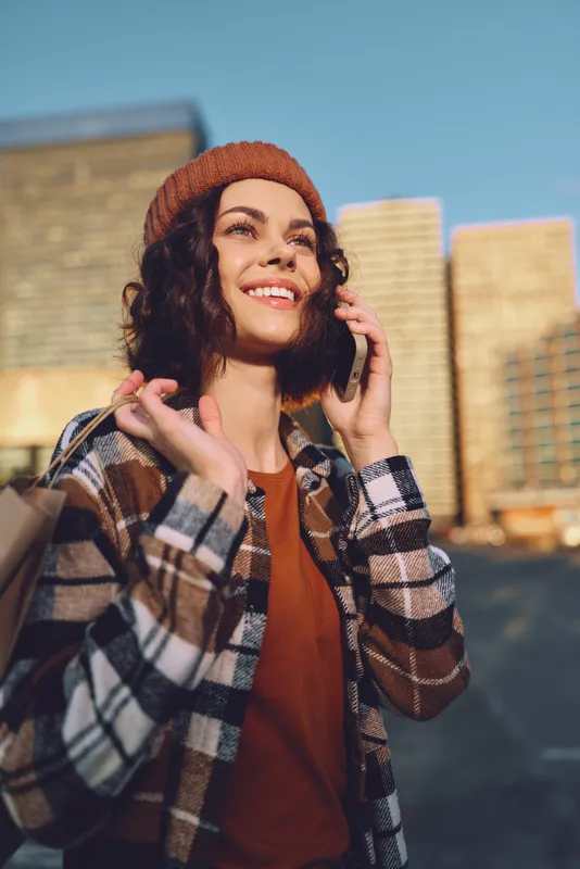 Woman on phone with smile in city during golden hour sunlight, holding shopping bag and wearing beanie and plaid jacket; authenticity, candid lifestyle, emotional storytelling mood.