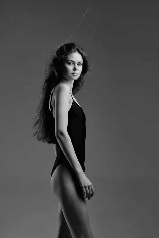 Black and white portrait of a slender woman in a bodysuit with long hair on a gray background.