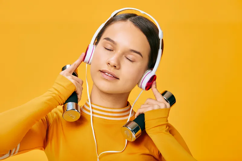 Woman fitness enthusiast wearing a bright orange sweater, headphones on, eyes closed, holding dumbbells for a focused workout session in a studio.