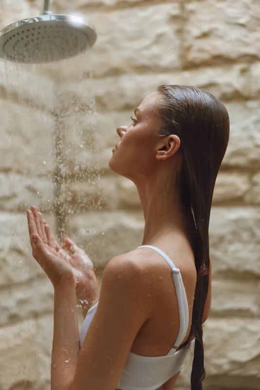 young woman enjoying warm shower water, wet hair, peaceful expression, natural stone wall background, intimate wellness moment
