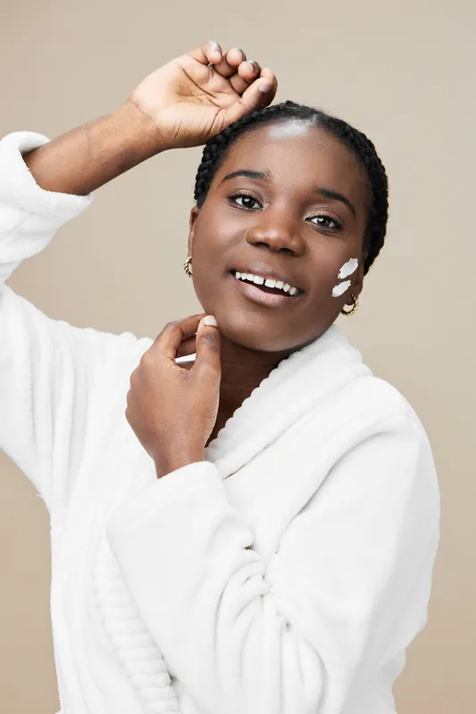 Smiling young woman applying moisturizer in a cozy white robe against a neutral background, showcasing a skincare routine for natural beauty, self care, and wellness