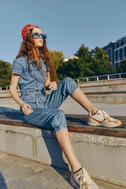 Lifestyle woman in modern boho-western denim street style sitting outdoors with sunglasses and a red bandana, captured in warm film color tones.