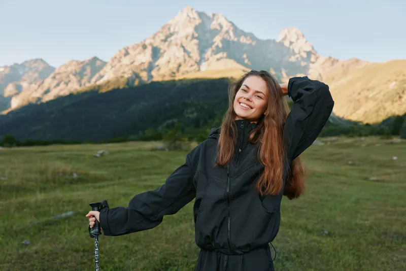 A cheerful woman stands outdoors in a scenic mountain meadow, smiling as she holds a trekking pole, enjoying fresh air and natural beauty around her.