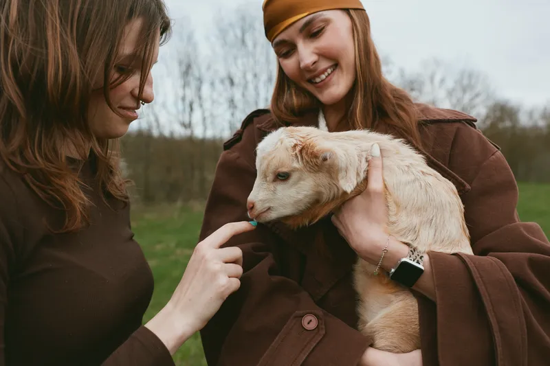 Two women in an outdoor setting share a tender moment with a small kid goat, one gently strokes the animal while the other smiles, conveying friendship in a casual lifestyle scene outdoors