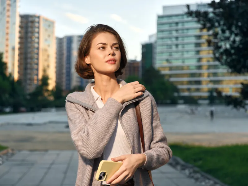 A cheerful business professional woman stands outdoors in a modern urban setting, texting on a smartphone, smiling confidently, projecting a positive, dynamic work vibe.
