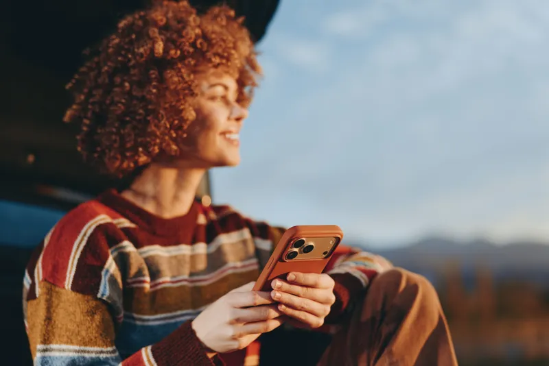 Happy woman lifestyle outdoors with curly hair holding smartphone in hands, warm sunset light, plaid sweater, side profile near car, communication and modern tech, relaxed travel mood