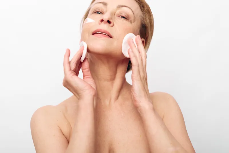 Mature woman with bare shoulders gently applies facial toner using cotton pads. Soft natural skincare routine concept focusing on cleansing, hydration, and anti-aging beauty treatment.