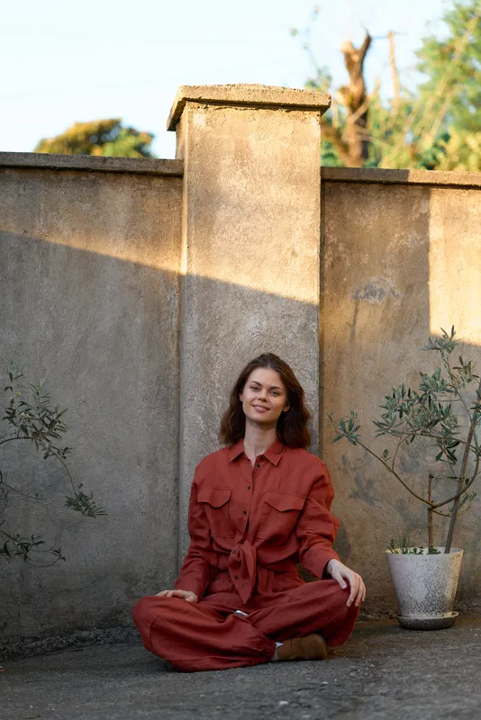 Cheerful woman outdoors, seated crosslegged, rust outfit, sunlit moment, relaxed posture, soft breeze, natural colors, lush greenery background