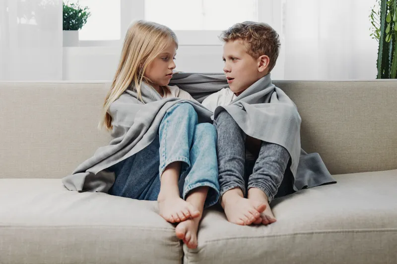 Two children wrapped in a grey blanket sitting barefoot on a beige couch, looking at each other in a bright living room with plants in the background.