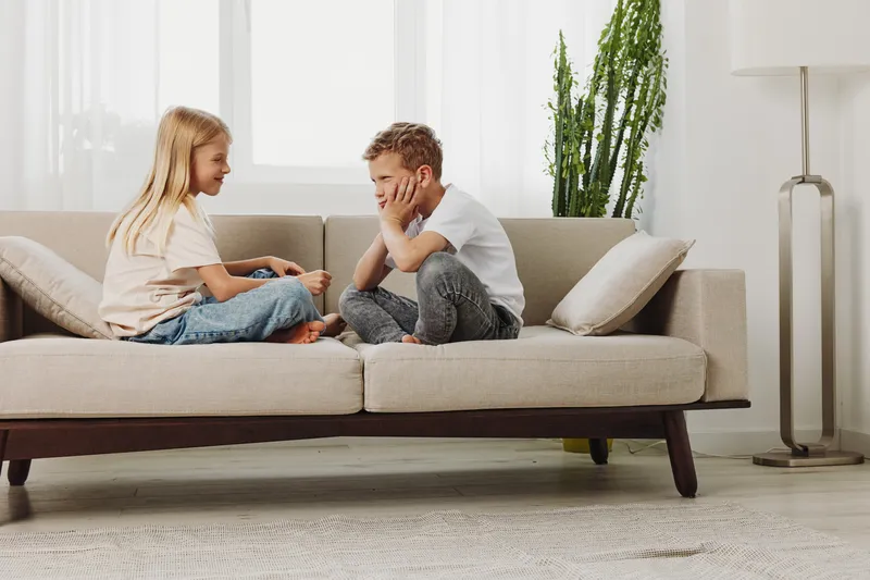 Two children sitting on beige sofa in bright room, talking and smiling, concept of childhood friendship and communication.