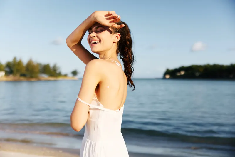 Sunlit portrait of a joyful woman on a sandy beach, wearing a white dress, shielding eyes with a hand, wind in hair, horizon over calm water.