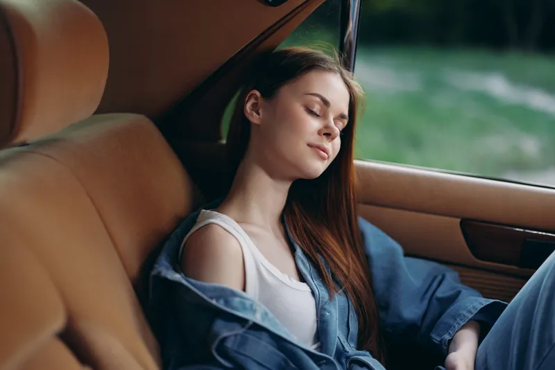 Young woman resting in a car with a serene expression, wearing a casual outfit, showcasing natural beauty and tranquility amid a blurred green background.