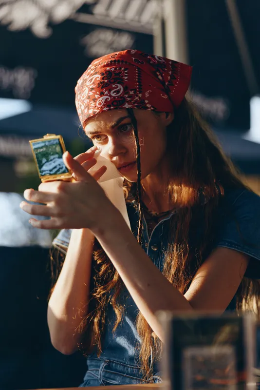 young woman with red bandana closely examining object in hand while sitting in cafe with warm natural light and casual denim outfit