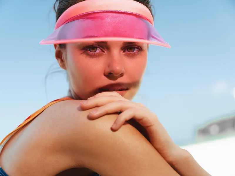 Close up portrait of a young woman wearing a pink translucent visor outdoors, sunlit skin, confident gaze, and relaxed pose with crossed arms.