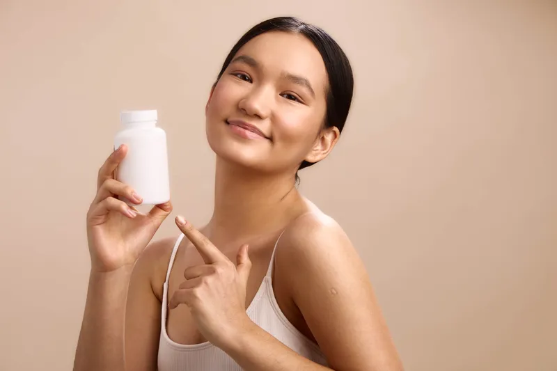 Asian woman holds a white bottle and smiles in a soft studio setting, presenting a skincare product with a friendly, confident vibe and clean, minimal aesthetics.