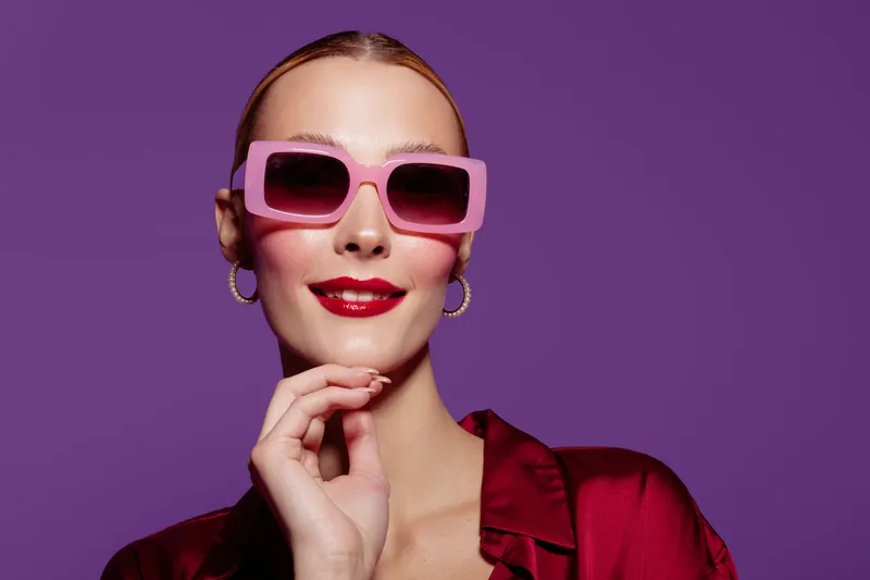 Stylish woman with pink sunglasses and red lipstick poses confidently on purple background. Fashion concept with bold makeup, accessories, and modern glamour style portrait.
