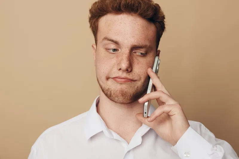 Young man with red hair and freckles wearing white shirt talks on smartphone with skeptical expression on beige background. Concept of communication, doubt, and casual lifestyle.