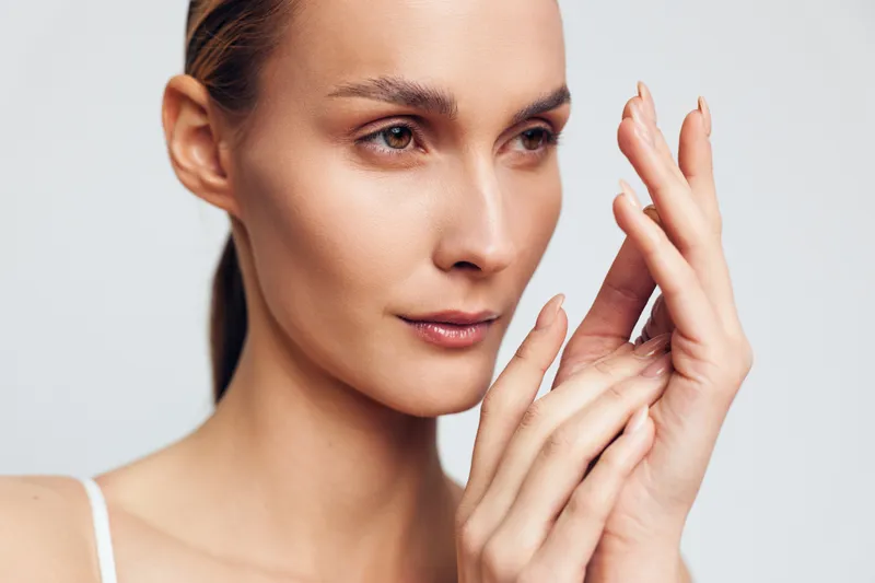 A close-up portrait of a young woman with clear skin gently touching her face and hands against a neutral background. Concept for skincare, beauty, and natural care.