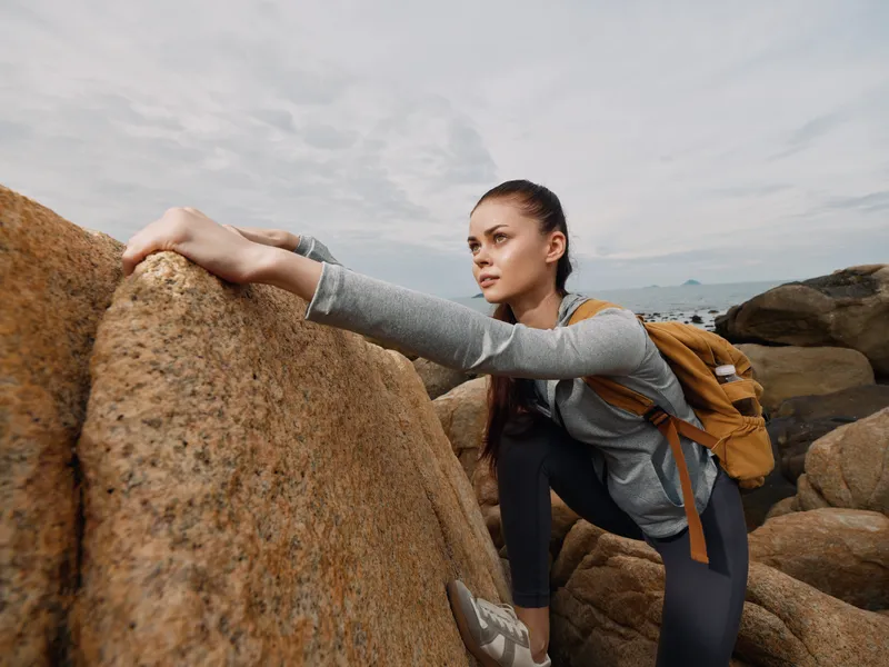 Young woman climbing on rocky terrain near the ocean, wearing sporty attire, expressing determination and focus, ideal for adventure and fitness themes
