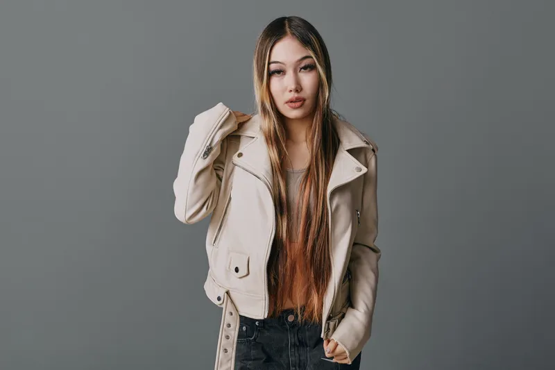 Young woman with long straight hair wearing beige jacket and black pants posing on grey background for fashion, beauty and casual style portrait photography.