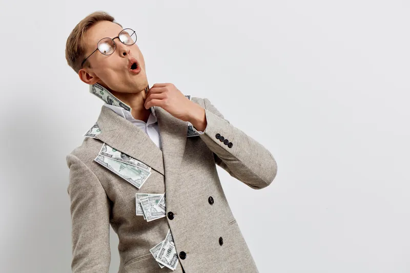 Businessman wearing glasses and suit with money bills attached to blazer, looking surprised and thinking, isolated on plain white background, studio portrait. Business success concept.
