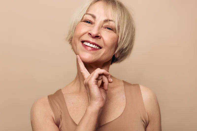 Smiling middle aged woman with short blonde hair touches chin gently. She wears beige tank top against beige background. Concept of natural beauty, confidence, and mature skincare.