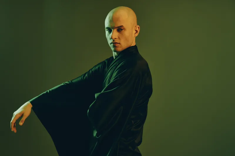 Handsome man in a black robe poses dramatically against a green backdrop, conveying confidence and artistry in his movements. Unique and captivating image.