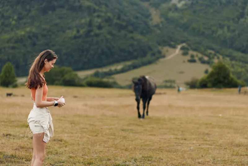 A casual woman stands in a sunlit meadow, watching a distant horse. She wears light clothing and a gentle smile, conveying calm, freedom, and a connection with nature.