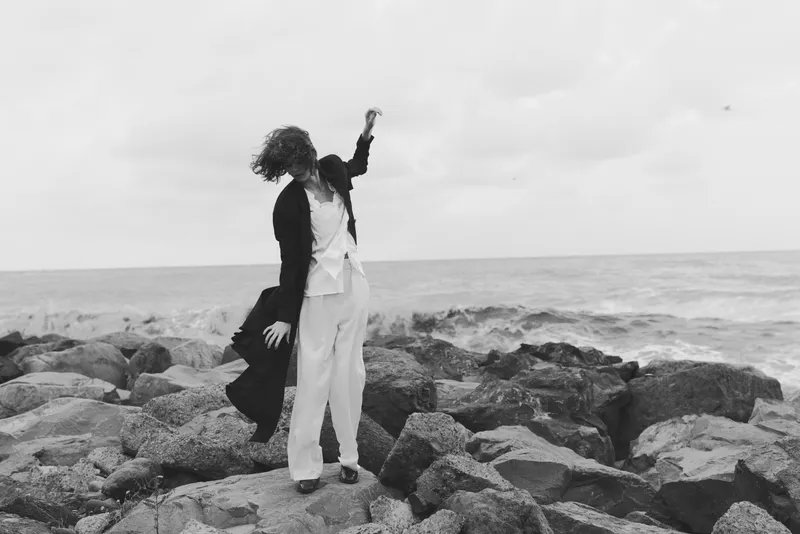 Monochrome scene of a woman standing on jagged rocks by the sea, wind blowing her coat as waves crash nearby, creating a dramatic, moody coastal moment with a sense of motion and freedom