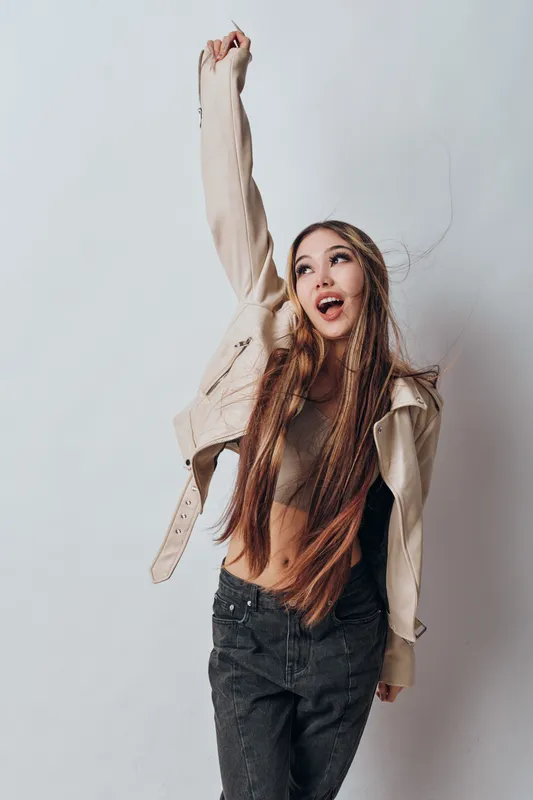 Young woman with long hair wearing casual jacket and jeans posing with raised hand and open mouth against plain background for fashion and lifestyle concept.