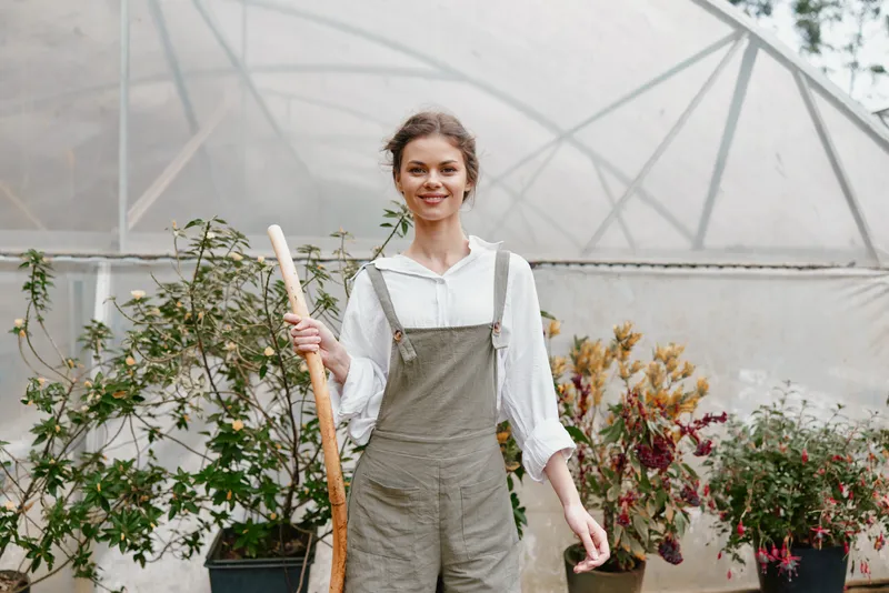 Young woman in a greenhouse wearing overalls, holding a gardening tool, showcasing a joyful expression amidst vibrant plants, representing sustainable gardening