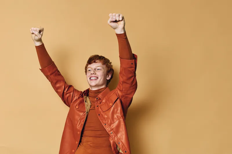 Happy young man with raised fists celebrating success in orange jacket and turtleneck against beige background. Smiling male expressing joy and victory indoors.